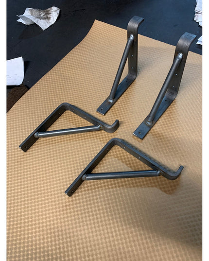 Four black metal Norwich Support Brackets by Industrial Farm Co displayed on textured surface, showing curved wrought iron...