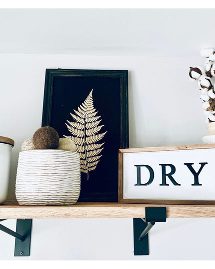 Black metal Nauseef shelf support brackets holding natural wood shelf with farmhouse decor including fern art and cotton s...