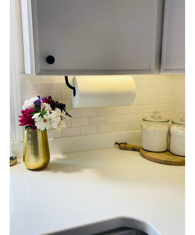 Black metal Hyde Hall Paper Towel Holder mounted under white kitchen cabinet with white paper towel roll on marble countertop