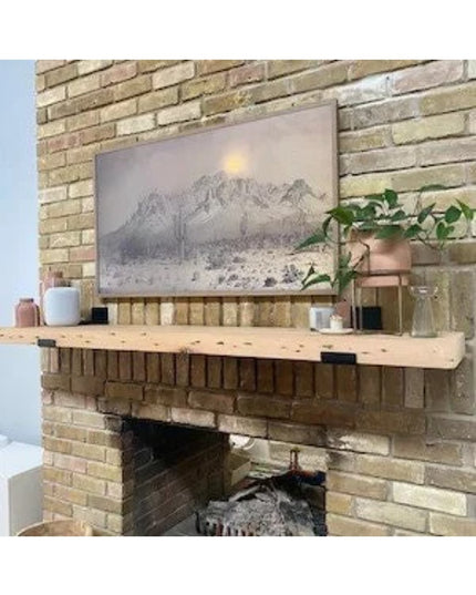 Rustic wooden floating shelf with metal brackets mounted on brick fireplace mantel displaying mountain artwork and decor i...