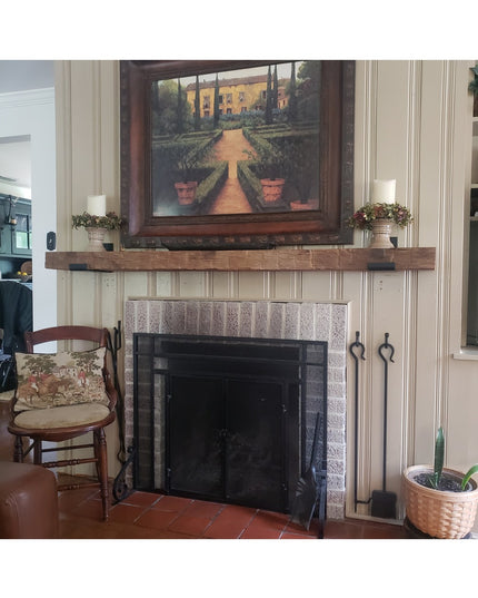 Rustic wooden mantel shelf with dark metal bracket supports mounted above stone fireplace in traditional living room setting