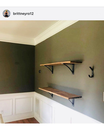 Two black steel Pops 2" industrial shelf brackets with curved support bars mounted on green wall holding natural wood shel...