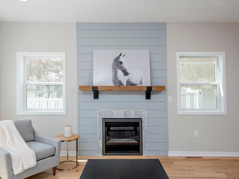 Blue-gray shiplap fireplace wall with honey-toned wood mantel held by black Kudlick farmhouse mantel brackets, featuring a black-and-white horse portrait, linen armchair, and gold side table