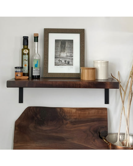 Dark walnut floating shelf with black steel L-brackets displaying kitchen items and framed artwork on white wall