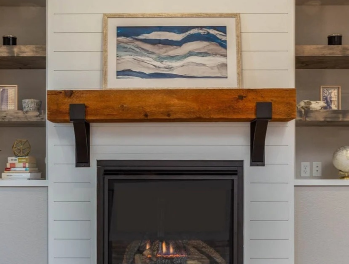 A rustic wood mantel with black metal brackets mounted above a fireplace, set against a white shiplap wall. A framed abstract landscape painting hangs above the mantel, and built-in side shelves on both sides display decorative items.