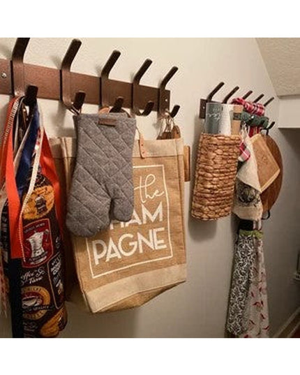 Wall-mounted wooden coat rack with multiple black metal hooks holding kitchen items including oven mitts, towels, and wick...