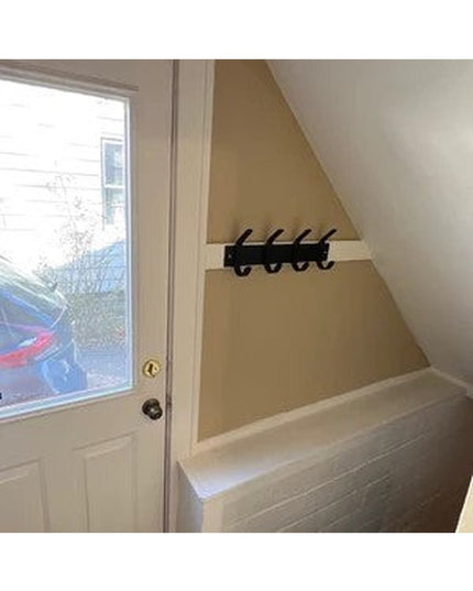 Black metal Fairmount Style Coat Rack with 4 hooks mounted on tan wall in entryway nook with white door and built-in bench