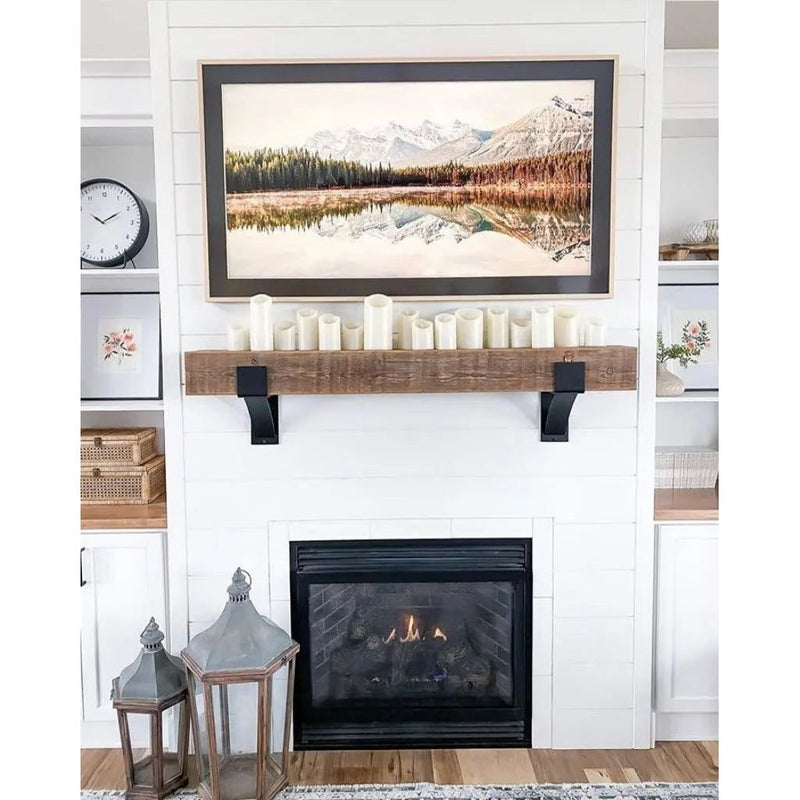 hite fireplace with reclaimed wood mantel held by black Kudlick farmhouse mantel brackets, styled with pillar candles, a framed mountain landscape, and galvanized lanterns on the hearth.