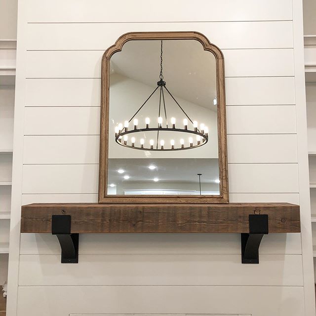 Chunky reclaimed wood mantel mounted on white shiplap wall with black Kudlick farmhouse mantel brackets, topped with an arched wood-framed mirror reflecting a black chandelier.