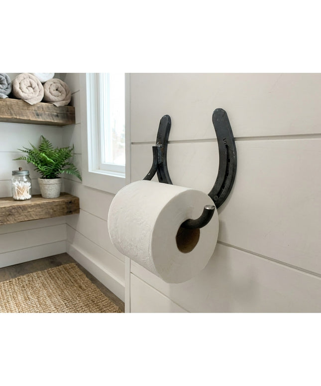 Rustic black horseshoe toilet paper holder mounted on white shiplap bathroom wall with farmhouse decor and floating wood s...